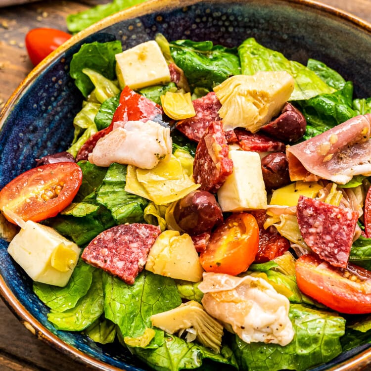 Portrait photo of a dark blue ceramic bowl with tossed Antipasto Salad in it on a wooden background