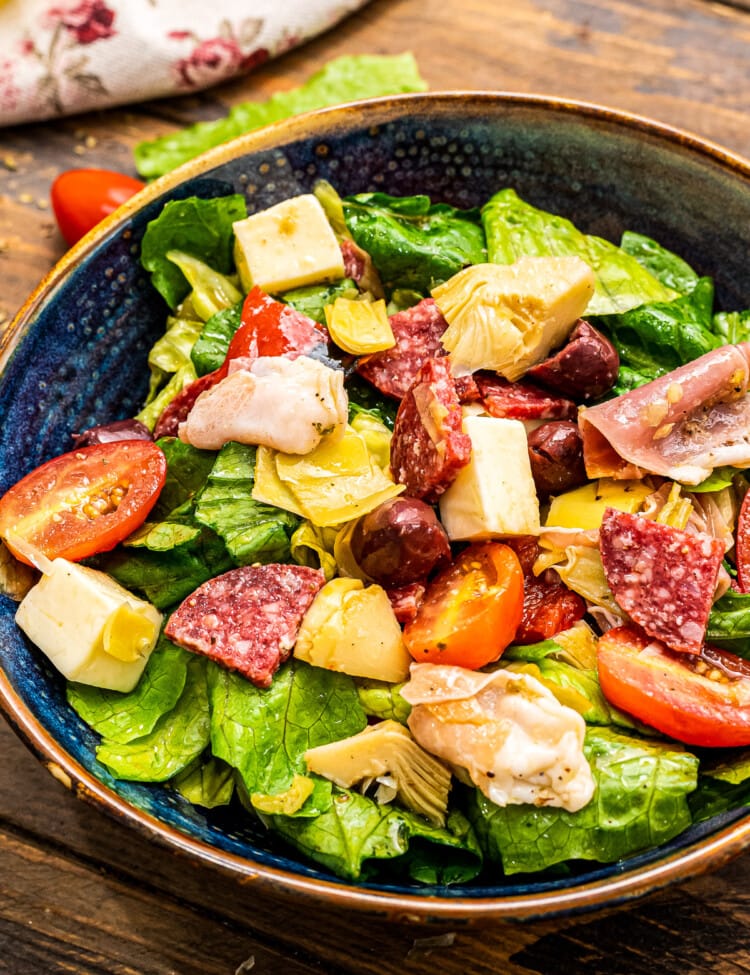 Portrait photo of a dark blue ceramic bowl with tossed Antipasto Salad in it on a wooden background