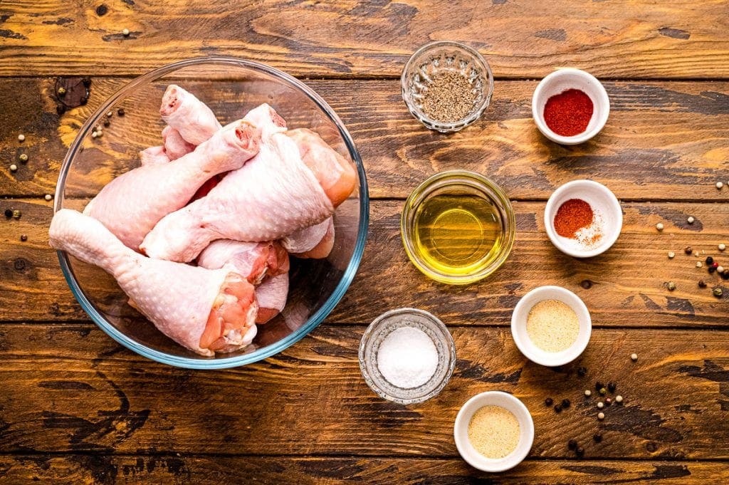 Overhead image of ingredients for baked chicken legs in glass bowls including chicken legs, oil, and seasonings.