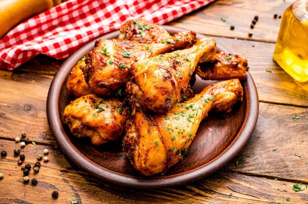 Wooden plate with a stack of chicken drumsticks garnished with chopped parsley with a red checkered napkin in background.