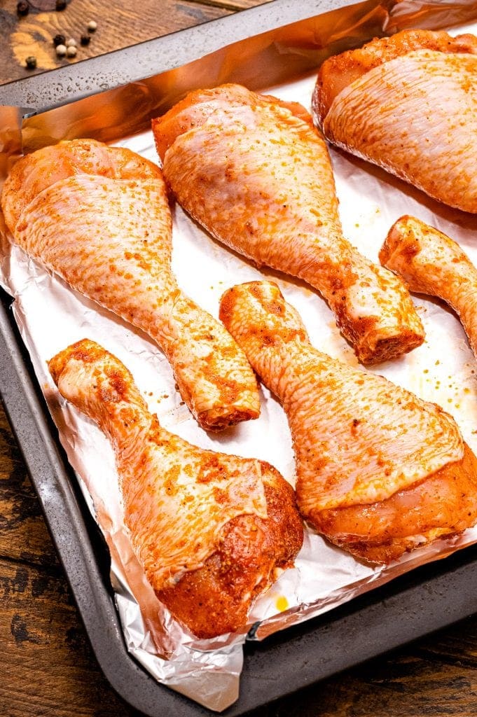 Raw chicken legs that are seasoned with seasonings and laying on aluminum foil lined baking sheet.