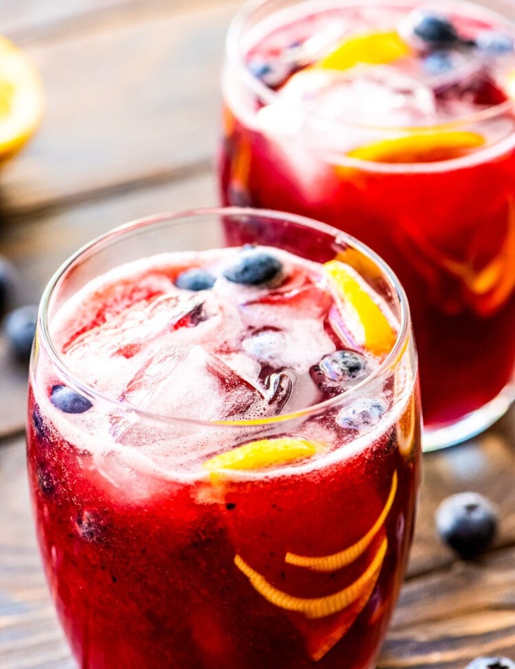 Two glasses on wooden background of freshly made blueberry lemonade with ice, blueberries, and lemon slices in glasses.