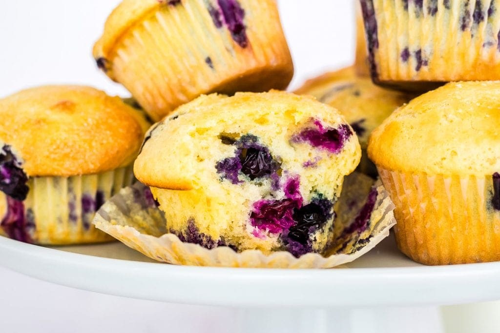 Blueberry muffin that is unwrapped and bite gone sitting on the wrapper with more muffins next to it