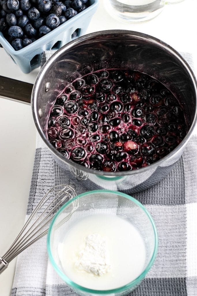 Bowl of water and cornstarch with a cooked blueberries in saucepan behind it.