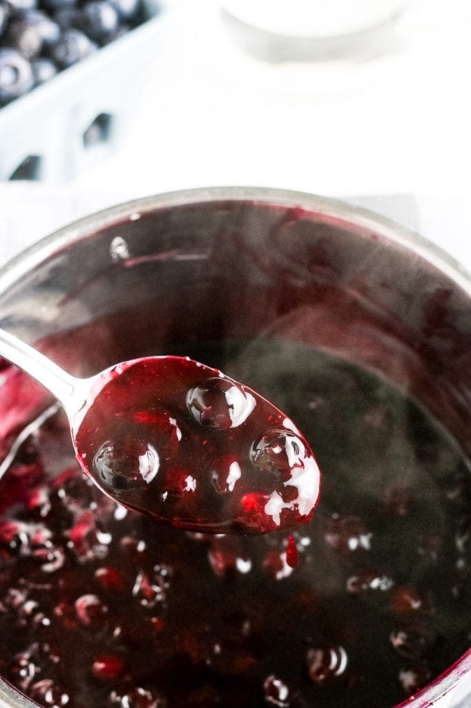 Spoon with fresh blueberry sauce on it. A pan of sauce in background