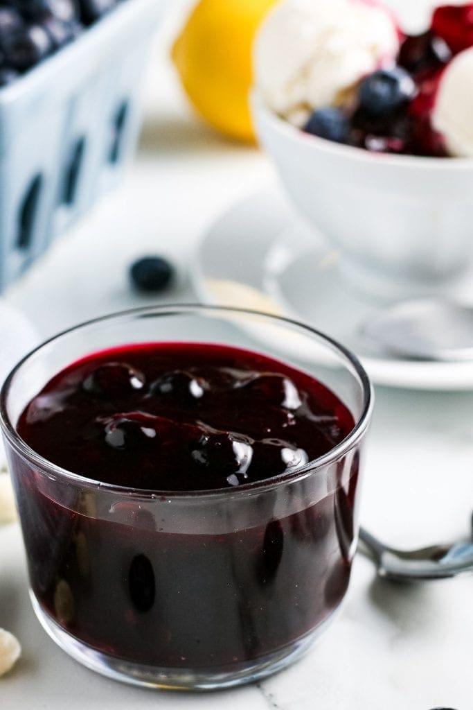 Glass dish full of easy blueberry sauce. Carton of fresh blueberries and bowl of vanilla ice cream with sauce on it in background.