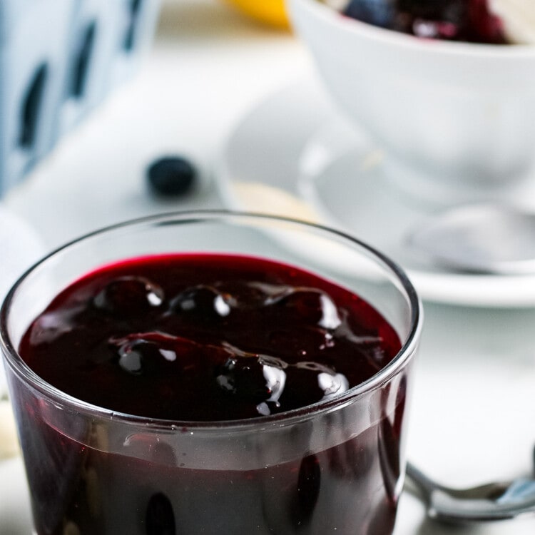 Glass dish full of easy blueberry sauce. Carton of fresh blueberries and bowl of vanilla ice cream with sauce on it in background.