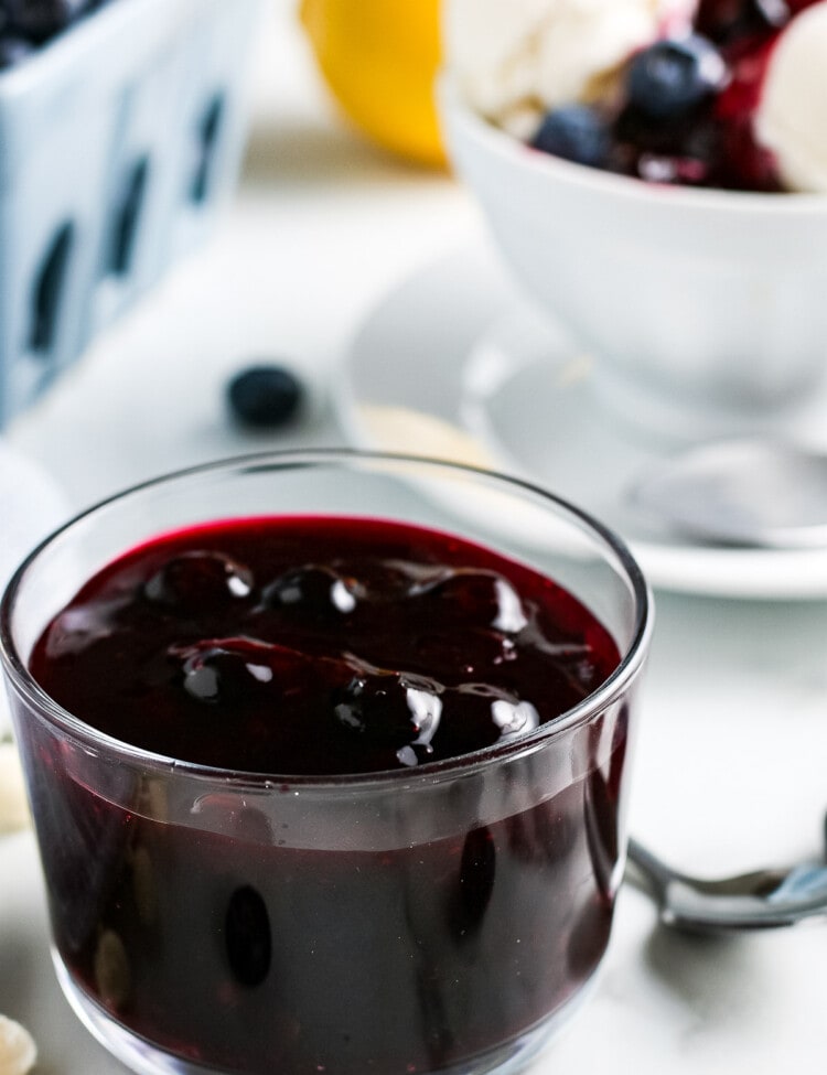 Glass dish full of easy blueberry sauce. Carton of fresh blueberries and bowl of vanilla ice cream with sauce on it in background.