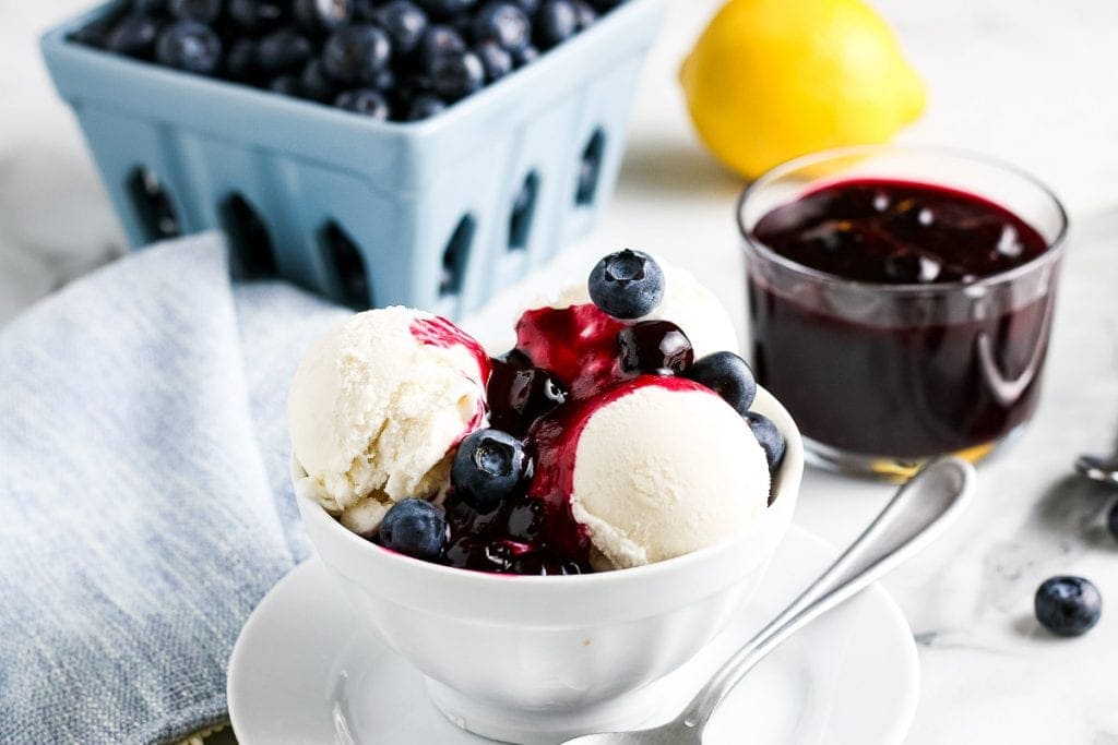 Vanilla ice cream in bowl topped with blueberry sauce. A bowl of sauce and fresh blueberries in the background.