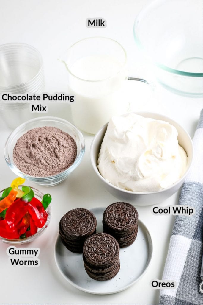 Ingredients to make Dirt Cups like white plate with Oreos, chocolate pudding mix in white bowl, a bowl of Cool Whip and a bowl of gummy worms.