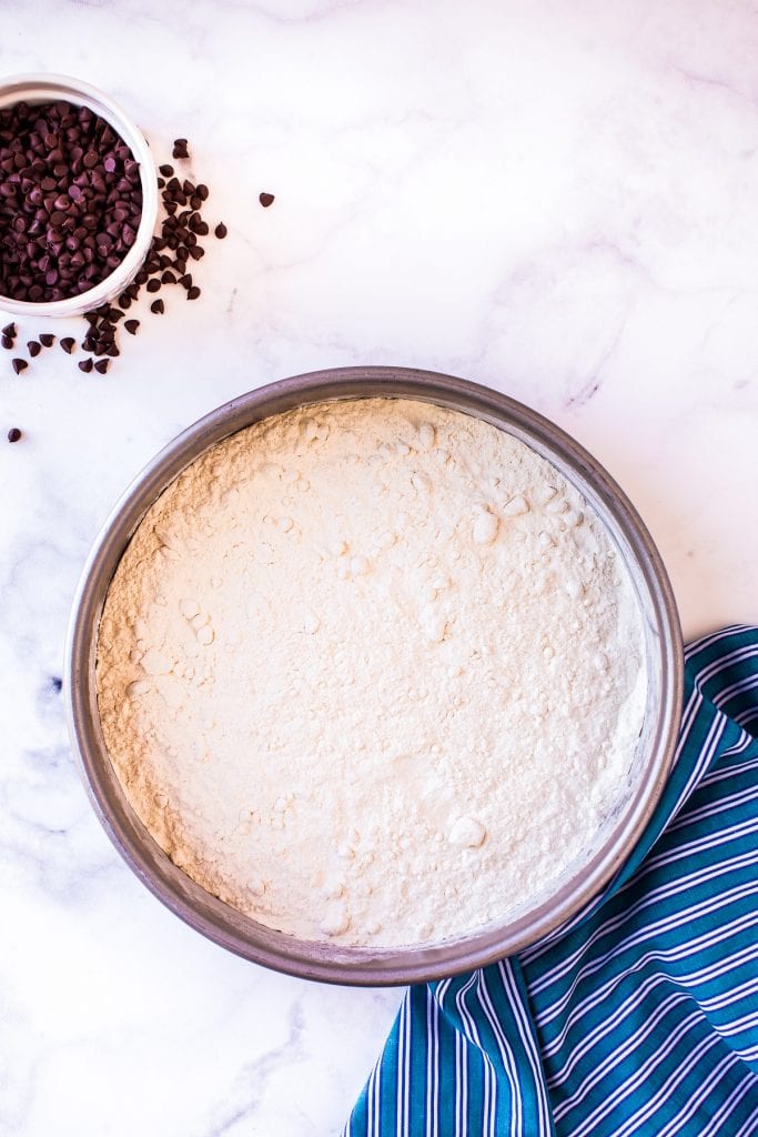 Overhead image of flour in bowl with blue napkin and chocolate chips beside it.