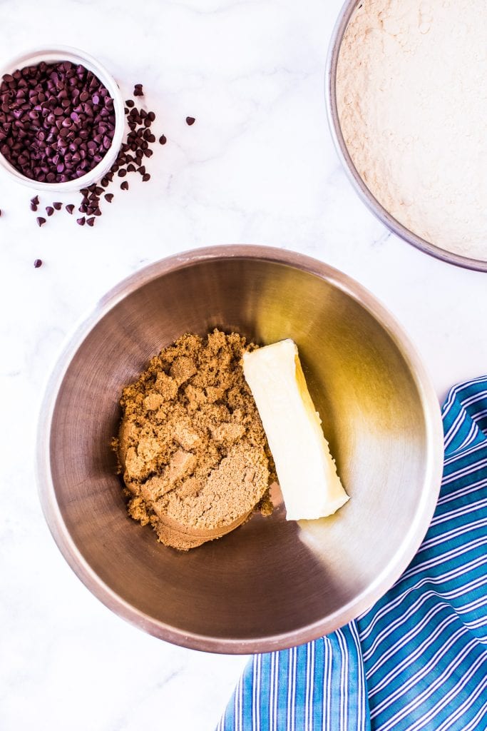 Overhead image of brown sugar and stick of butter in bowl with blue napkin and chocolate chips beside it.