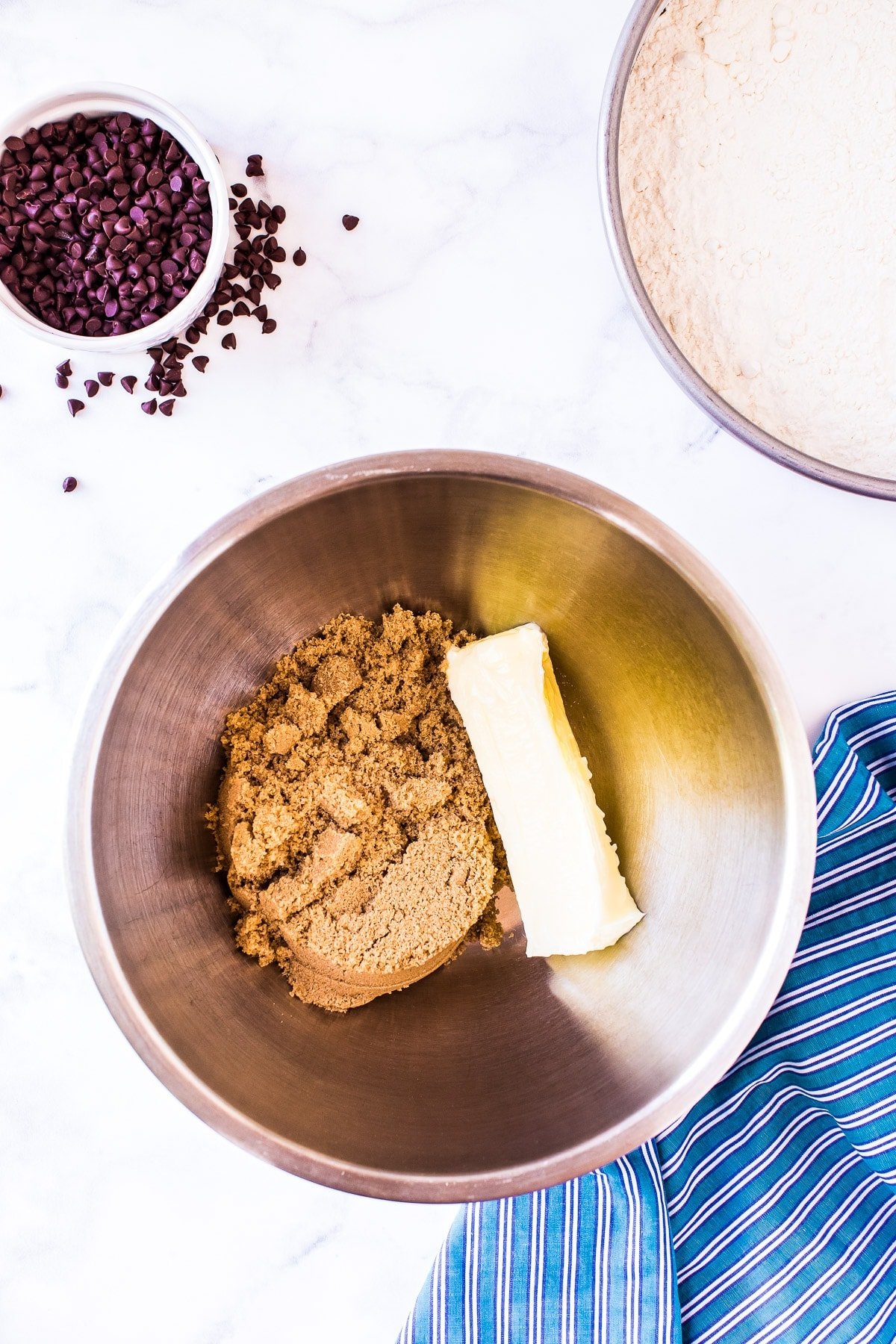 Overhead image of brown sugar and stick of butter in bowl with blue napkin and chocolate chips beside it.