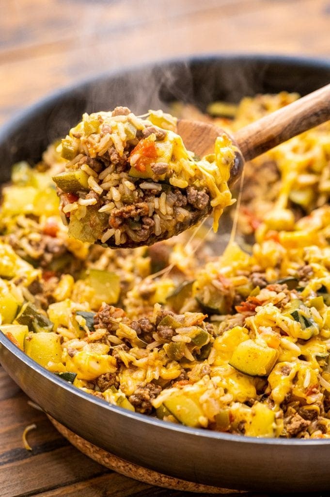 Wooden spoon scooping ground beef and zucchini skillet out of pan with steam coming off of it.