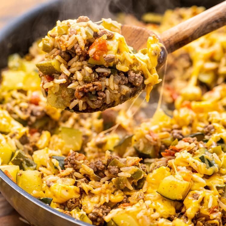 Wooden spoon scooping ground beef and zucchini skillet out of pan with steam coming off of it.