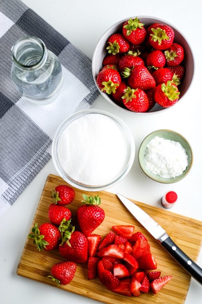 Wood cutting board with chopped strawberries on it along with the rest of the ingredients in background for strawberry sauce.