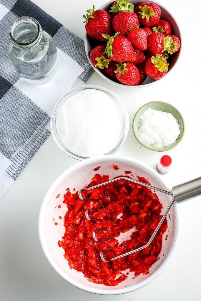White bowl with mashed strawberries in it along with the rest of the ingredients in background for strawberry sauce.