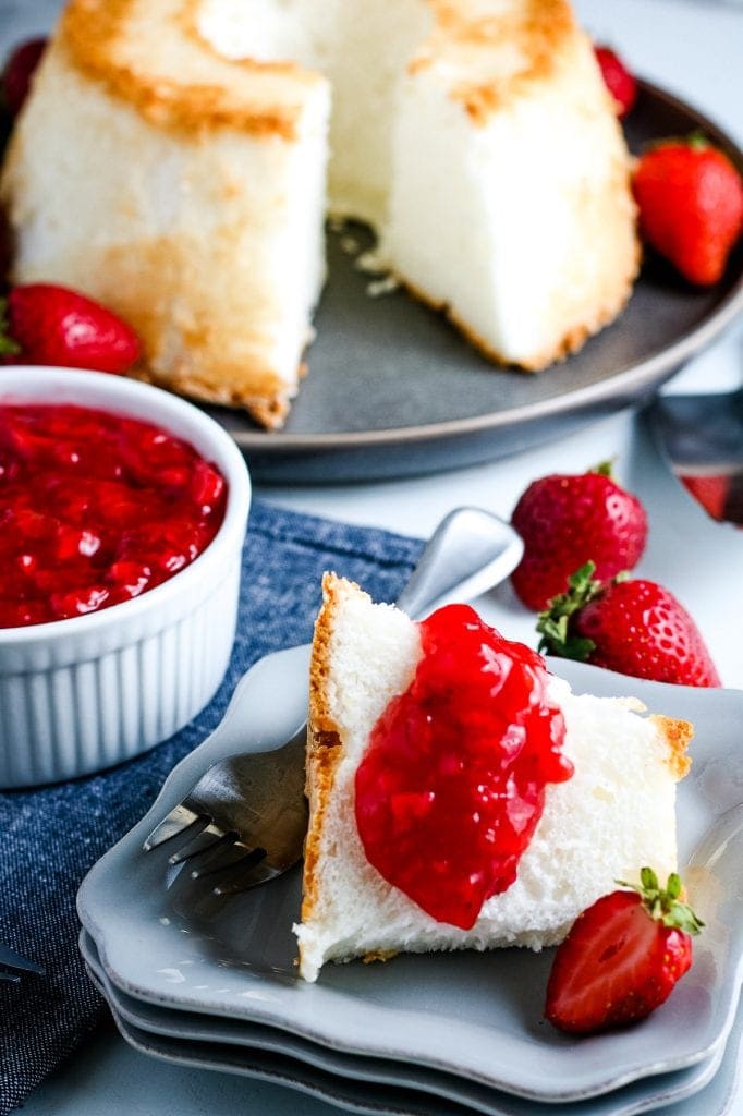 Slice of Angel food cake on plate with strawberry sauce. An angel food cake in background with strawberry sauce in dish.