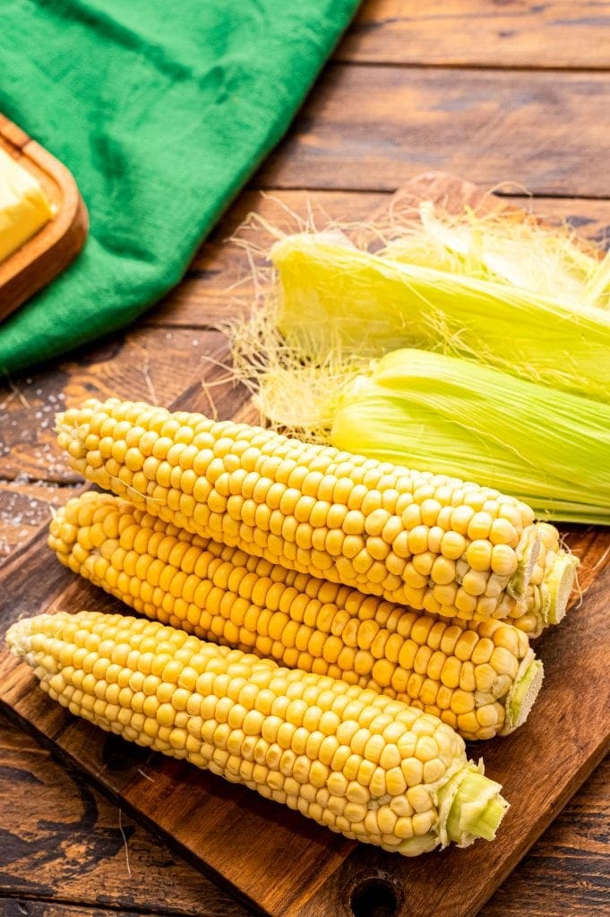 Four pieces of corn on the cob on a wood cutting board with husk behind them.