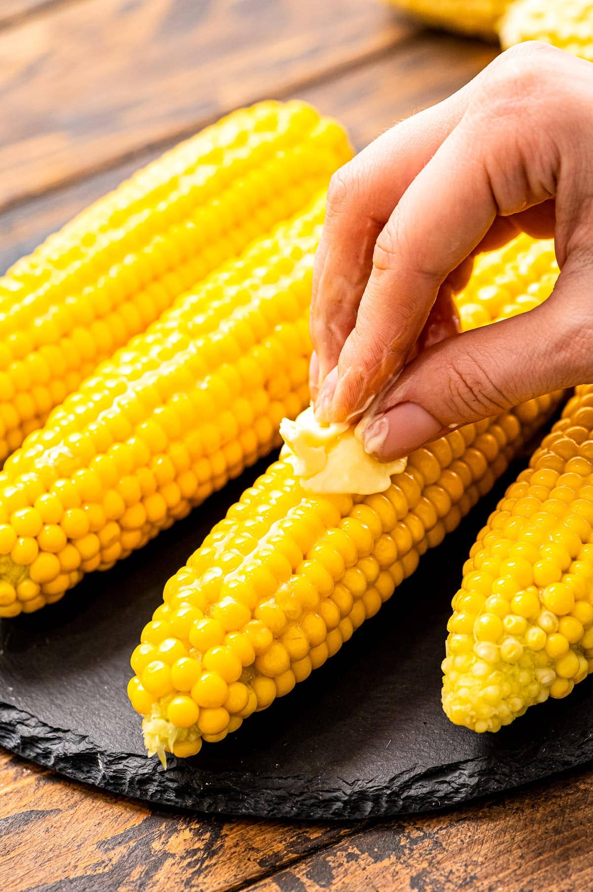 Hand rubbing a slice of butter over a piece of corn on the cob.