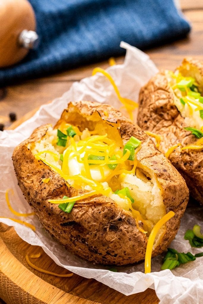 Baked Potatoes on freezer paper on a brown wooden plate. Baked Potatoes are cut open showing the fluffy inside and topped with cheese and green onion.