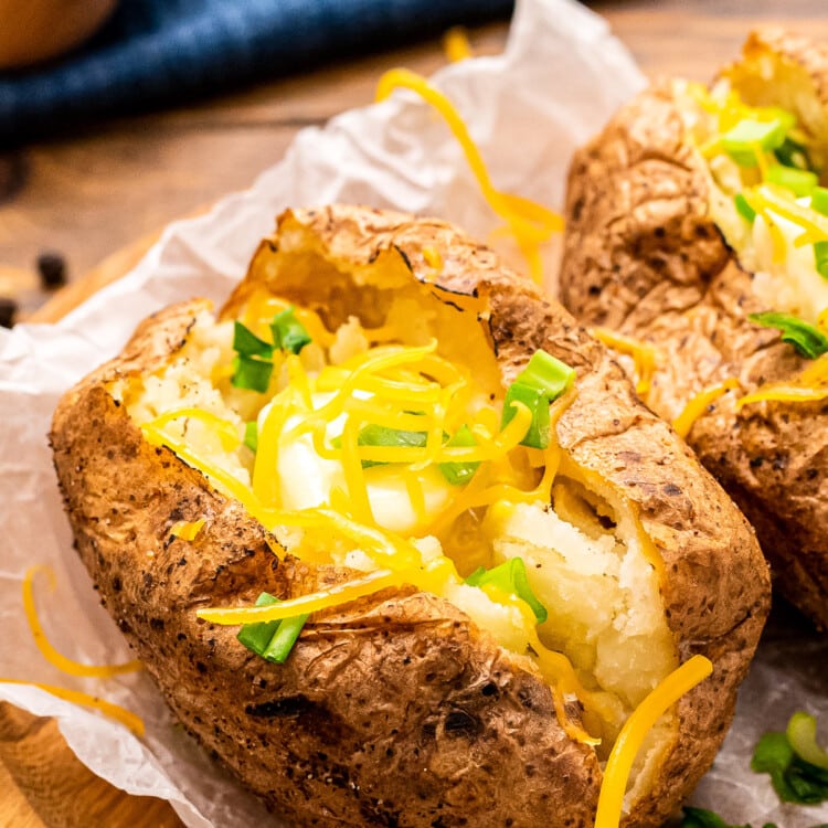 Baked Potatoes on freezer paper on a brown wooden plate. Baked Potatoes are cut open showing the fluffy inside and topped with cheese and green onion.