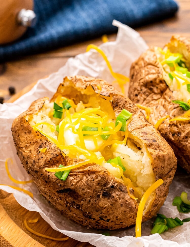 Baked Potatoes on freezer paper on a brown wooden plate. Baked Potatoes are cut open showing the fluffy inside and topped with cheese and green onion.