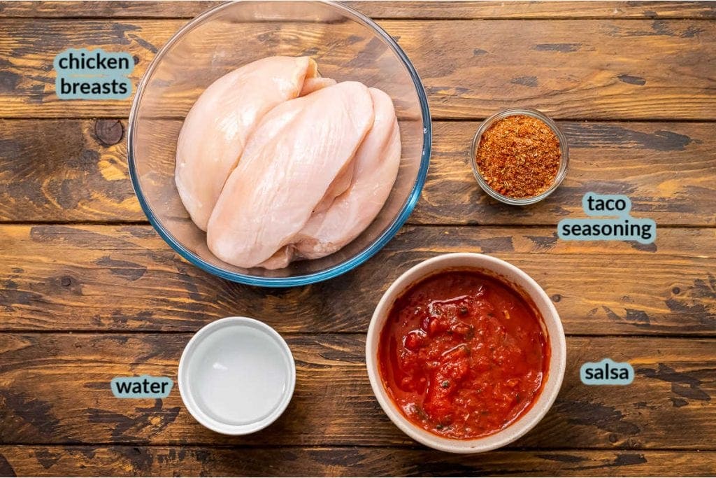 Wooden background with glass bowls with raw chicken breasts, water, salsa and taco seasoning