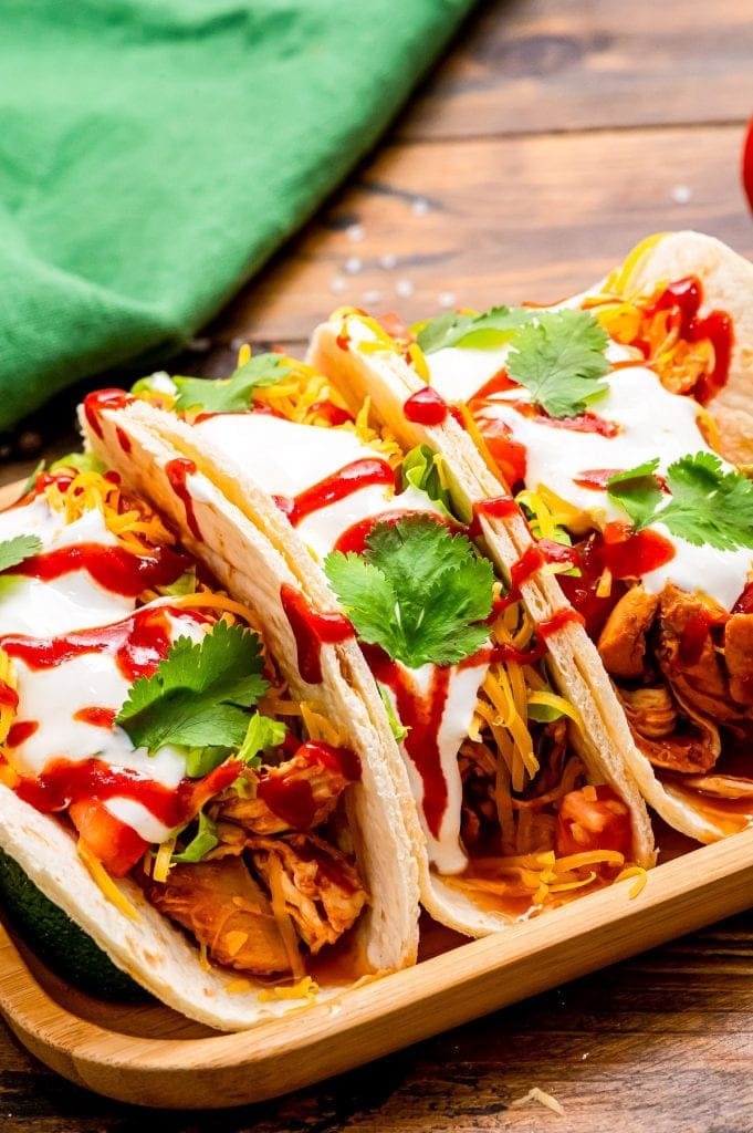 Wooden serving platter with three tacos on top of it. Tacos filled with shredded chicken, shredded cheese, diced tomatoes, csour cream, cilantro and hot sauce with green napkin in background