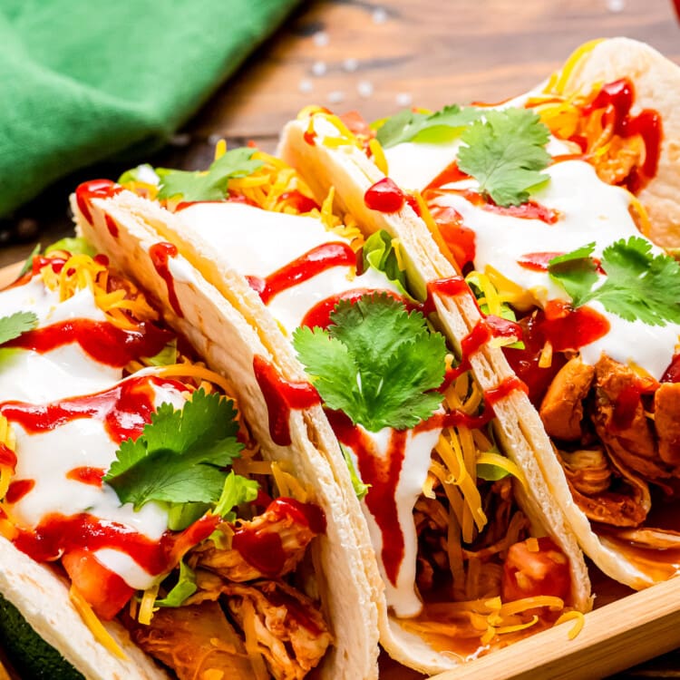 Wooden serving platter with three tacos on top of it. Tacos filled with shredded chicken, shredded cheese, diced tomatoes, csour cream, cilantro and hot sauce with green napkin in background