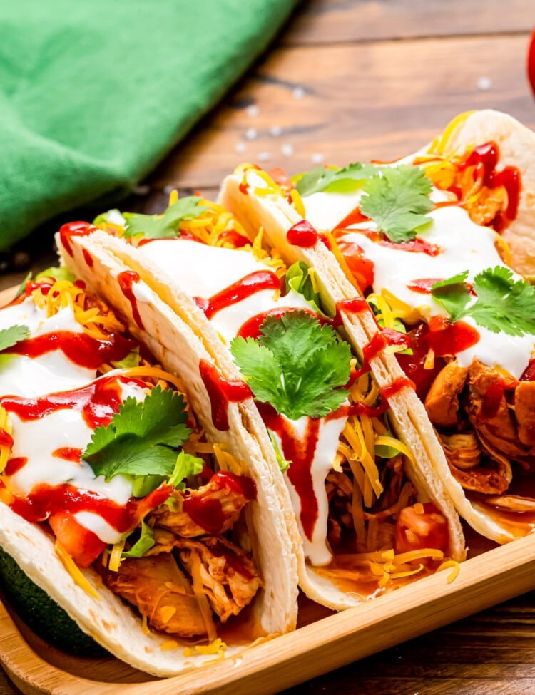 Wooden serving platter with three tacos on top of it. Tacos filled with shredded chicken, shredded cheese, diced tomatoes, csour cream, cilantro and hot sauce with green napkin in background