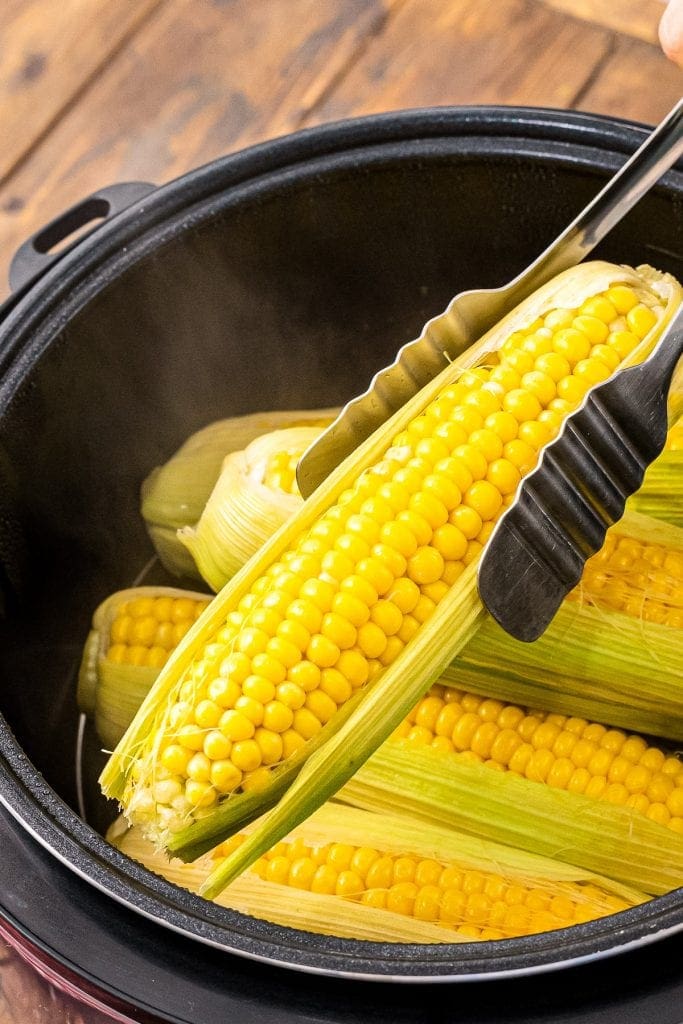 Tongs lifting a piece of corn on the cob with husk on out of Instant Pot.