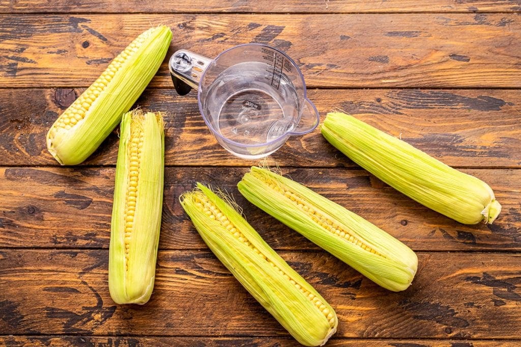 Overhead image of corn on the cob in husks and a cup of water.