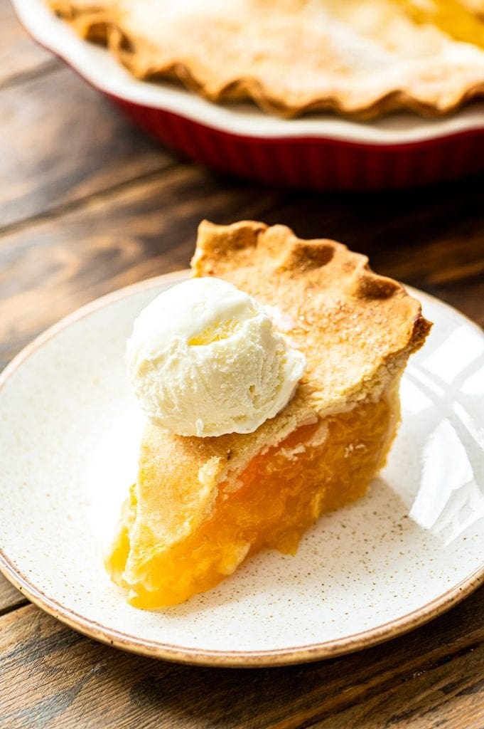 Slice of peach pie topped with ice cream on a light colored plate. Full peach pie in background