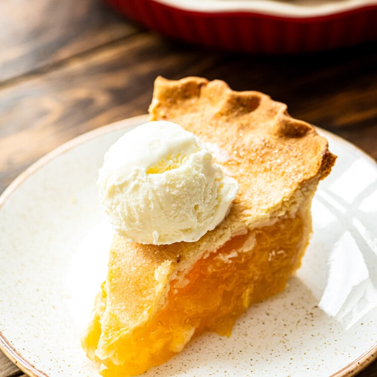 Slice of peach pie topped with ice cream on a light colored plate. Full peach pie in background