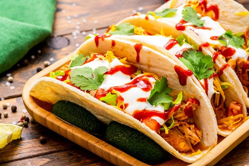Landscape picture of Wooden serving platter with three tacos on top of it. Tacos filled with shredded chicken, shredded cheese, diced tomatoes, csour cream, cilantro and hot sauce with green napkin in background