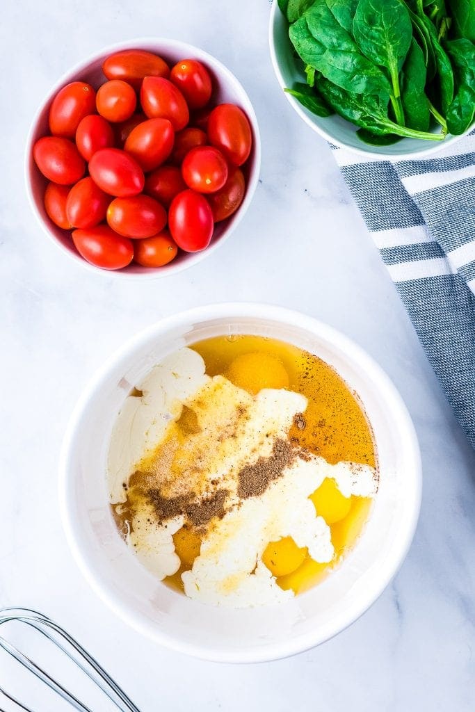 White bowl with eggs, heavy cream, garlic powder, onion powder and nutmeg, Bowl of cherry tomatoes and spinach beside it.