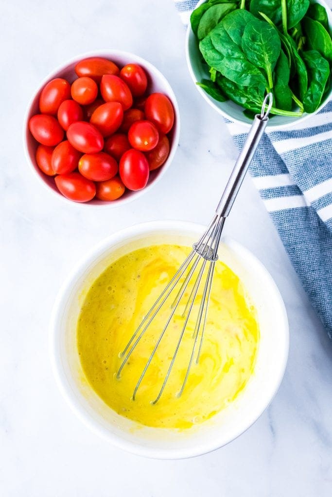 White bowl with whisked eggs, bowl of cherry tomatoes and a bowl of spinach on white background