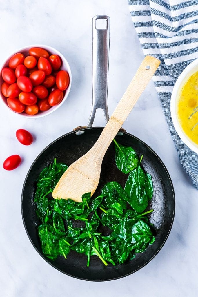 Cast Iron skillet with wilted spinach, a bowl of cherry tomatoes on light background