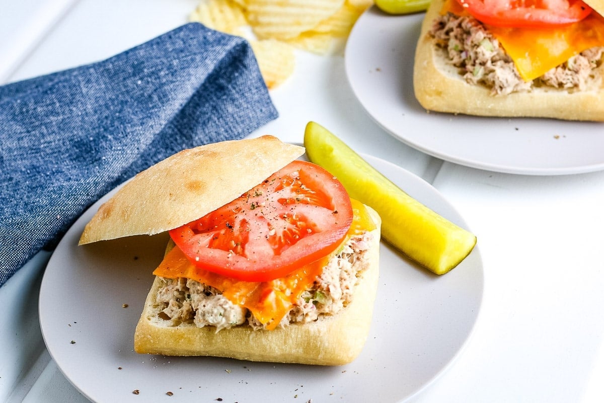 A light colored plate with a tuna melt sandwich on top with pickle next to it. A blue napkin and another plate with tuna melt on it in background