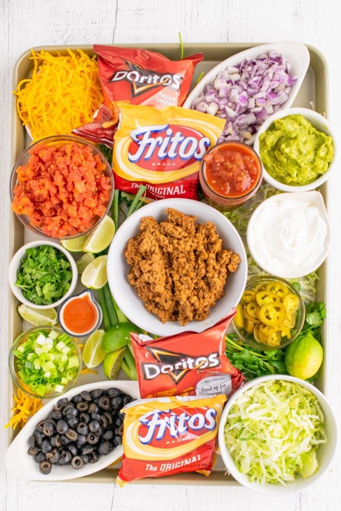 Overhead photo of sheet pan with bowls of taco toppings in bowl along with tomato, green onion to garnish. Small bowls of salsa and guacamole added plus small bags of Fritos and Nacho Cheese Doritos.