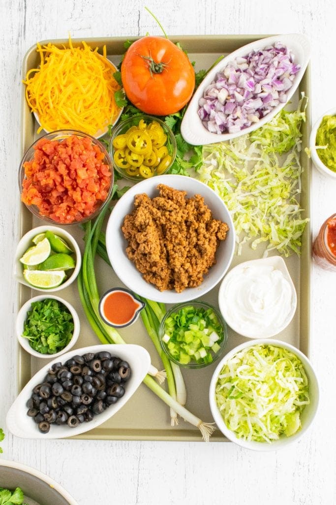 Overhead photo of sheet pan with bowls of taco toppings in bowl along with tomato, green onion to garnish