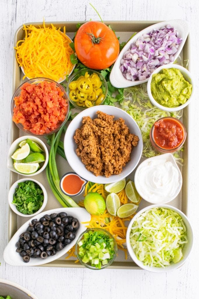 Overhead photo of sheet pan with bowls of taco toppings in bowl along with tomato, green onion to garnish. Small bowls of salsa and guacamole added.