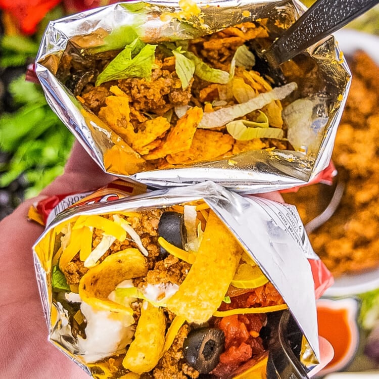 Hand holding two Walking Tacos. One made with Fritos and one made with Nacho Cheese Doritos.