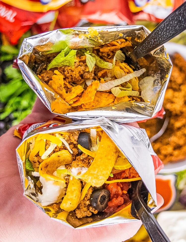 Hand holding two Walking Tacos. One made with Fritos and one made with Nacho Cheese Doritos.