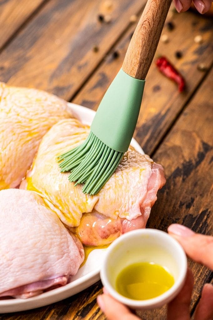 Brush coating chicken thigh with oil on a white plate