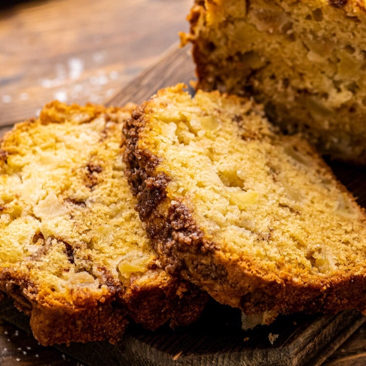 Two slices of apple bread next to the rest of the loaf on wood cutting board.