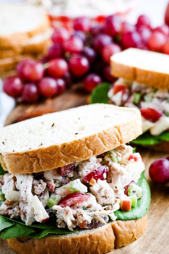 Close up image of sandwich filled with chicken salad and lettuce. Grapes and another sandwich in background.