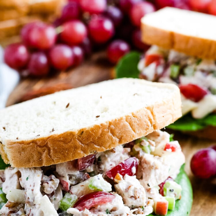 Close up image of sandwich filled with chicken salad and lettuce. Grapes and another sandwich in background.