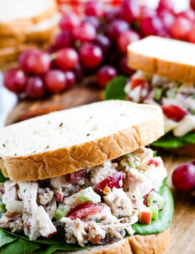 Close up image of sandwich filled with chicken salad and lettuce. Grapes and another sandwich in background.